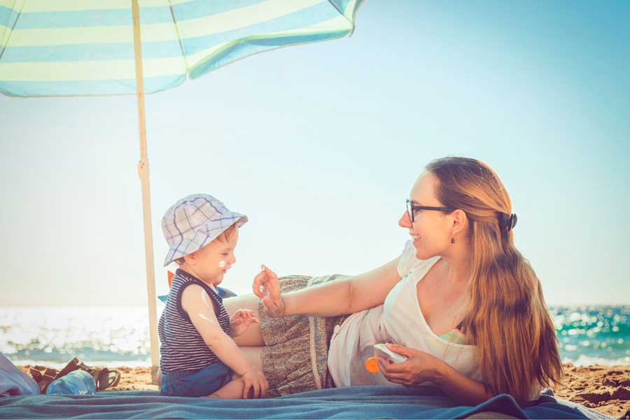 Mutter cremt ihr Kleinkind unter einem Sonnenschirm am Strand mit Sonnencreme ein